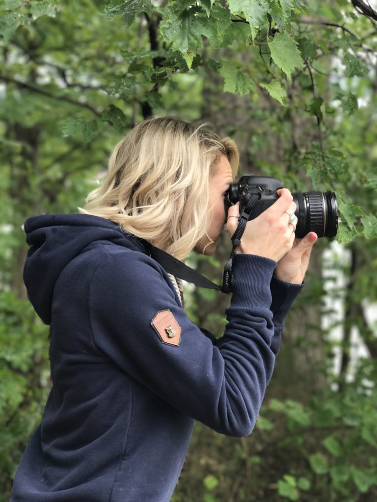 joelle holding camera up to her face taking a photo in a navy blue hoodie in the woods. 