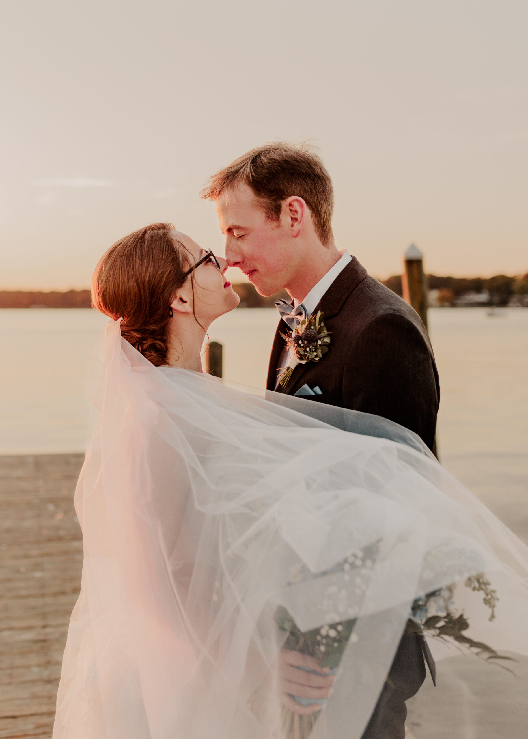 bride and groom facing each other, very close, soft smiles, brides veil blowing across their bodies in the wind with calm waterfront behind them